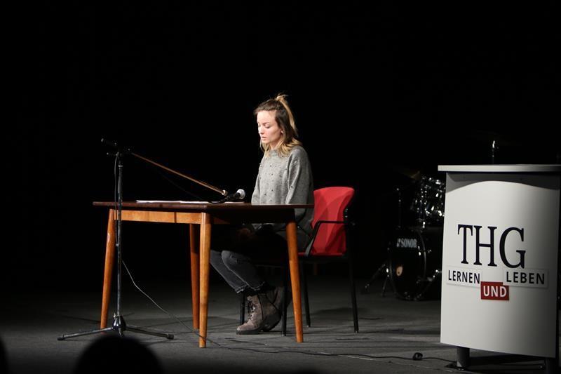 Eine Frau sitzt an einem Tisch auf der Bühne und liest aus den Protokollen vor.